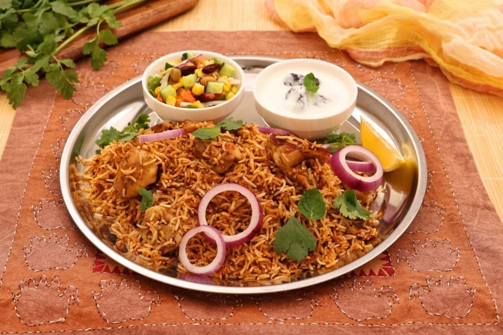 Biryani thali with raita and salad on a steel plate with herbs and onion garnish